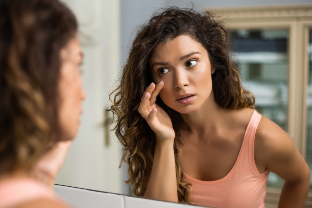 Mulher se olhando no espelho e analisando os olhos inchados para ilustração que a menstruação também pode causar olhos inchados. 