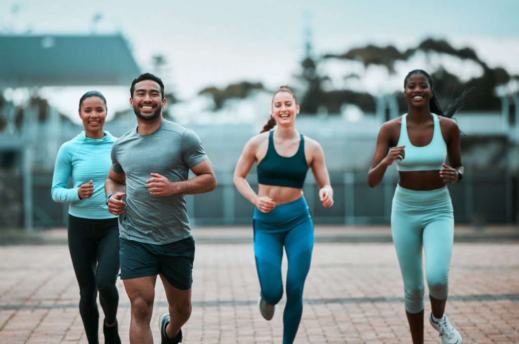 A imagem representa um grupo de 4 pessoas, duas mulheres negras, uma mulher branca, um homem negro. Todos estão vestidos com roupas de praticar exercicios físicos. O grupo está correndo ao ar livre, todos estão sorrindo.
